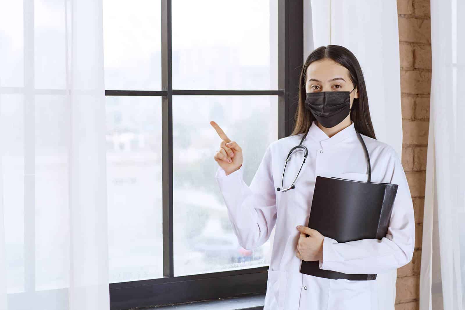 doctor with stethoscope black mask standing window holding black history folder patients while pointing somewhere
