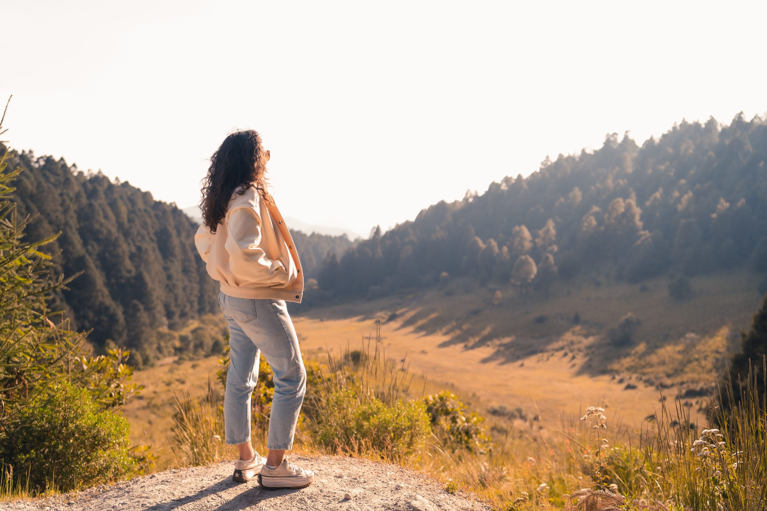 Woman standing on a dirt trail overlooking a valley with pine trees, wearing a peach jacket and jeans, facing away from the camera.