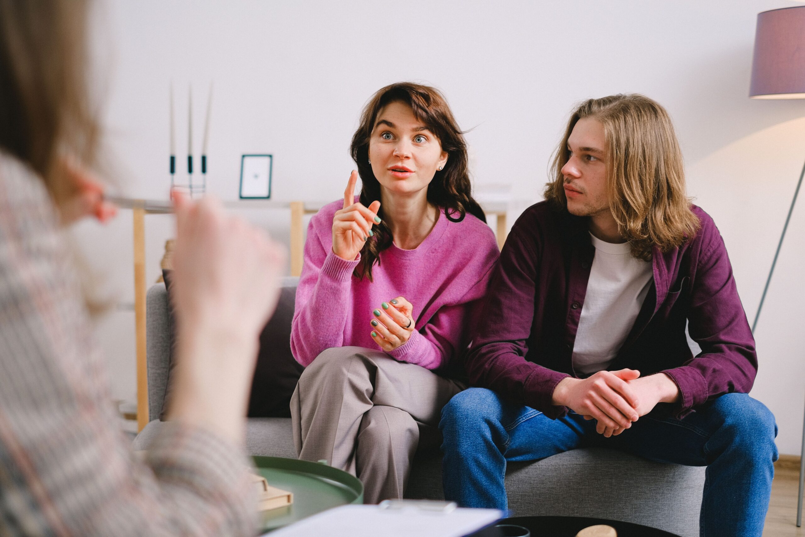 People sitting on a couch having a discussion in a cozy living room, woman in pink gesturing as she talks, man beside her listening.
