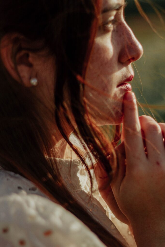 Close-up of a woman in profile with a finger pressed to her lips, signaling quiet, in warm outdoor light.