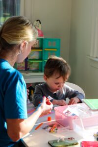 Adult woman in a blue shirt assisting a child with arts and crafts at a table full of colorful supplies in a bright room.