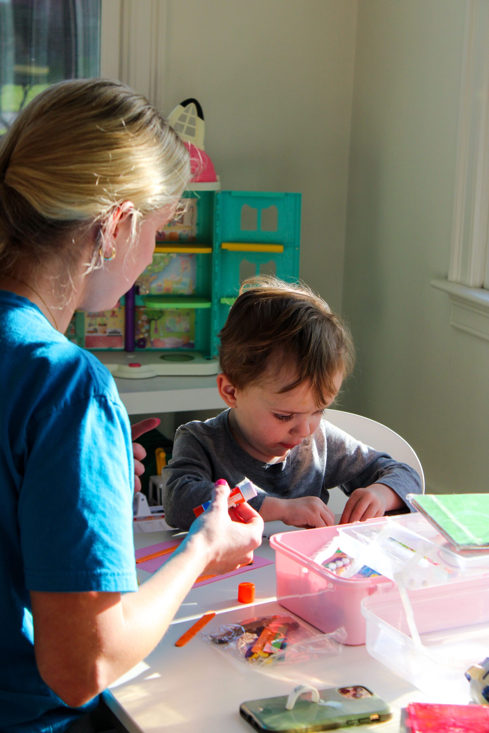 Adult woman in a blue shirt assisting a child with arts and crafts at a table full of colorful supplies in a bright room.