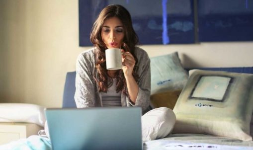 woman-in-grey-jacket-sits-on-bed-uses-grey-laptop-935743-740x493